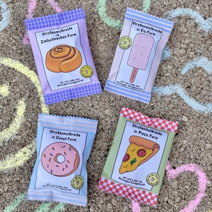 May include: Four colorful packages of sidewalk chalk shaped like different foods. The packages are labeled "Stra&szlig;enmalkreide" which is German for sidewalk chalk. The food shapes are a cinnamon roll, a popsicle, a donut, and a pizza.