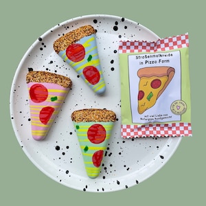 Op de afbeelding: Drie kleurrijke pizza-vormige stoepkrijtjes op een wit bord met zwarte stippen. De krijtjes zijn versierd met glitter en hebben rode, gele en groene toppings. De verpakking van de krijtjes is zichtbaar op de achtergrond en zegt "Stra&szlig;enmalkreide in Pizza-Form" en "mit viel Liebe aus Naturgips handgemacht!"