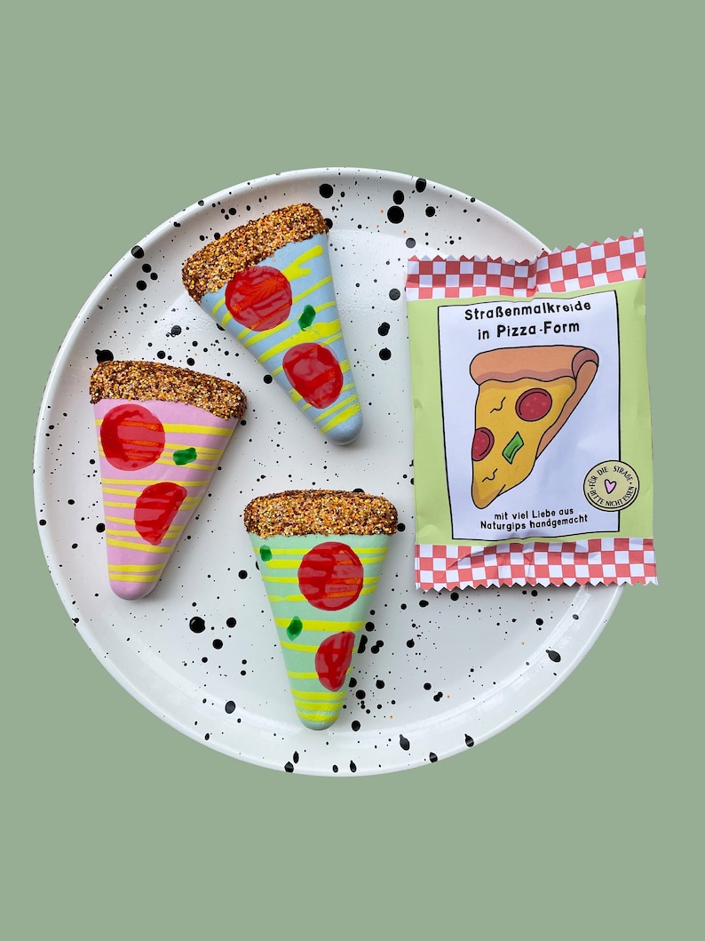 Op de afbeelding: Drie kleurrijke pizza-vormige stoepkrijtjes op een wit bord met zwarte stippen. De krijtjes zijn versierd met glitter en hebben rode, gele en groene toppings. De verpakking van de krijtjes is zichtbaar op de achtergrond en zegt "Stra&szlig;enmalkreide in Pizza-Form" en "mit viel Liebe aus Naturgips handgemacht!"