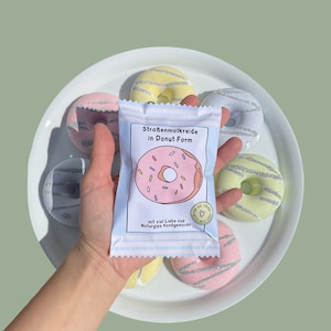 May include: A package of sidewalk chalk shaped like donuts. The package is white with pink and blue stripes and features a pink donut with sprinkles. The text on the package reads "Stra&szlig;enmalkreide in Donut-Form" and "mit viel Liebe aus Naturgips handgemacht!"