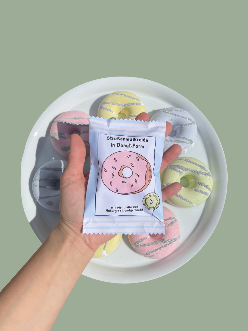 May include: A package of sidewalk chalk shaped like donuts. The package is white with pink and blue stripes and features a pink donut with sprinkles. The text on the package reads "Stra&szlig;enmalkreide in Donut-Form" and "mit viel Liebe aus Naturgips handgemacht!"