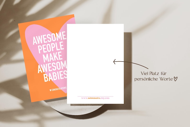 May include: A greeting card with the text "AWESOME PEOPLE MAKE AWESOME BABIES" in white on a pink heart shape, set against an orange background. A blank white card is next to it, with a website address at the bottom.