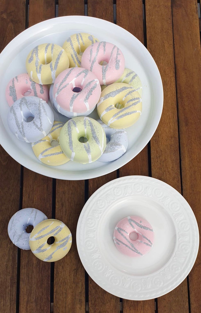 May include: A white plate with a variety of pastel-colored donuts with silver glitter. The donuts are arranged in a circle with the silver glitter facing up. The donuts are on a wooden table.
