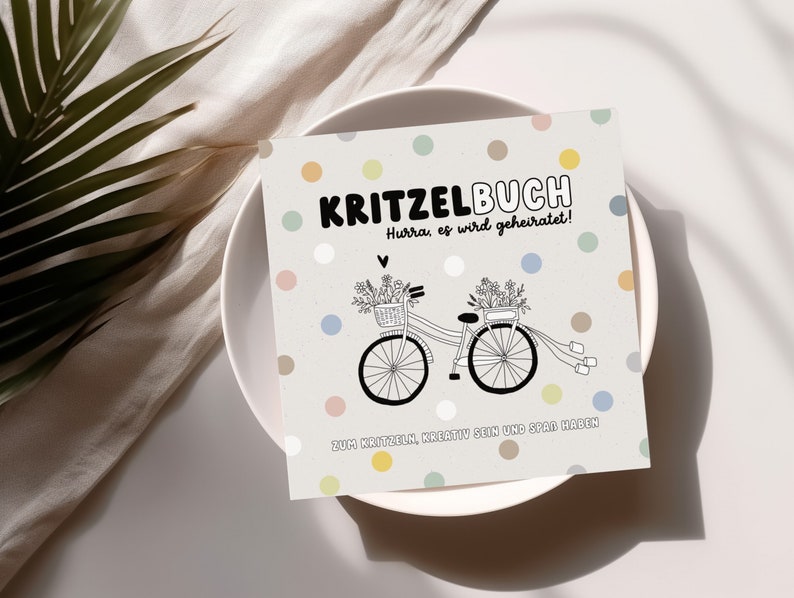 May include: A white card with a colorful polka dot pattern. The card has black text that reads "KRITZELBUCH Hurra, es wird geheiratet!" and "ZUM KRITZELN, KREATIV SEIN UND SPAR HABEN". There is a black and white illustration of a bicycle with flowers in baskets on the handlebars.