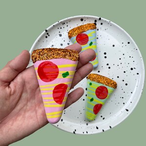 Op de afbeelding: Drie keramische pizza-puntversieringen met glitteraccenten. De punten zijn roze, blauw en groen met rode en gele toppings.