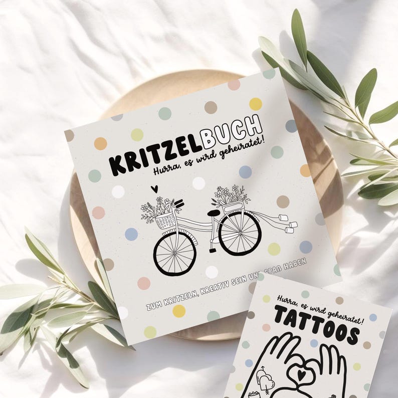 May include: A white card with a black and white illustration of a bicycle with flowers in the basket. The text on the card reads "Kritzelbuch Hurra, es wird geheiratet! Zum Kritzeln, kreativ sein und Spa&szlig; haben".