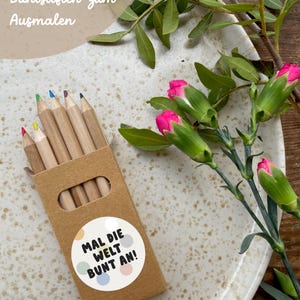 May include: A set of six wooden colored pencils in a brown cardboard box with a sticker that says "Mal die Welt Bunt an!"  The box is sitting on a white speckled plate with pink flowers.