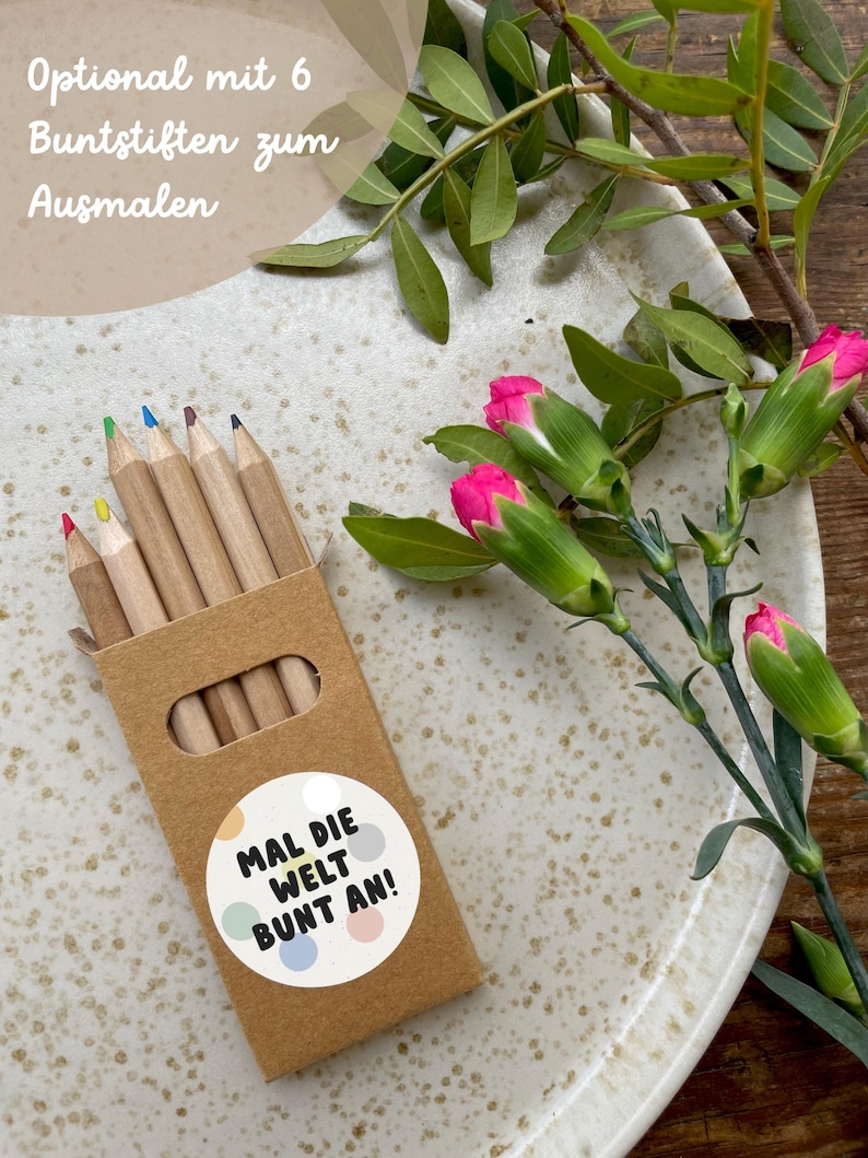 May include: A set of six wooden colored pencils in a brown cardboard box with a sticker that says "Mal die Welt Bunt an!"  The box is sitting on a white speckled plate with pink flowers.
