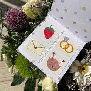 May include: A white card with a polka dot pattern on the back. The front of the card has four colourful temporary tattoos. The tattoos are a strawberry, a ring, an envelope with a heart, and a cartoon animal with hearts on its head. The card is surrounded by a bouquet of flowers.