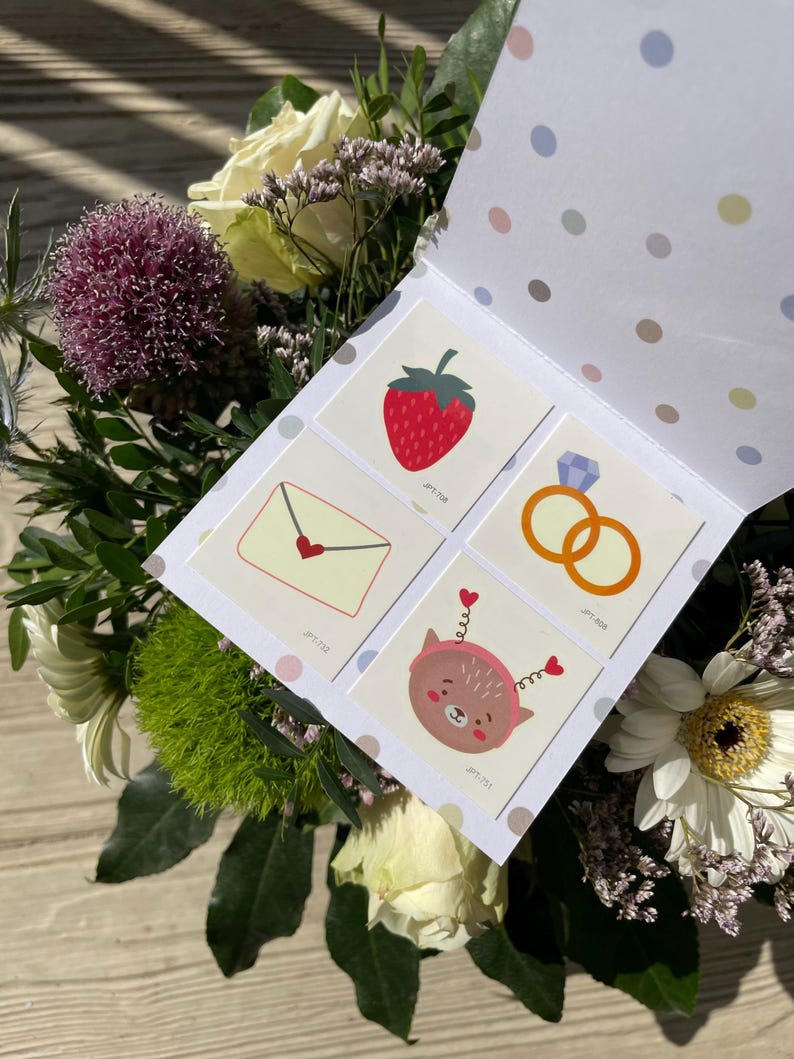 May include: A white card with a polka dot pattern on the back. The front of the card has four colourful temporary tattoos. The tattoos are a strawberry, a ring, an envelope with a heart, and a cartoon animal with hearts on its head. The card is surrounded by a bouquet of flowers.