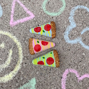 Op de afbeelding: Drie beschilderde pizzapunten met rode, gele en groene toppings en gouden glitteraccenten op een grijze achtergrond met krijttekeningen van geometrische vormen.