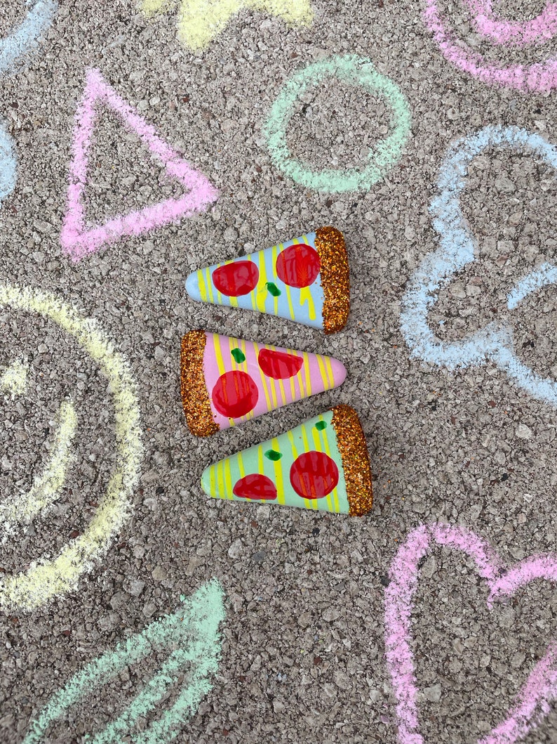 Op de afbeelding: Drie beschilderde pizzapunten met rode, gele en groene toppings en gouden glitteraccenten op een grijze achtergrond met krijttekeningen van geometrische vormen.