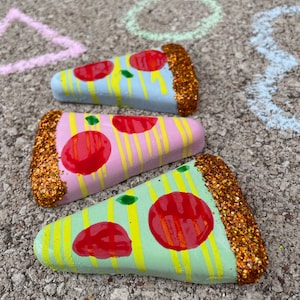 Op de afbeelding: Drie beschilderde pizzastukjes van steen met glitter. De pizzastukjes zijn in verschillende kleuren geschilderd, waaronder roze, blauw en groen. Elk stukje heeft rode pepperoni en gele en witte strepen.