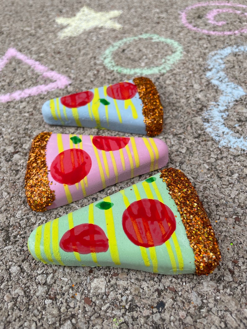 Op de afbeelding: Drie beschilderde pizzastukjes van steen met glitter. De pizzastukjes zijn in verschillende kleuren geschilderd, waaronder roze, blauw en groen. Elk stukje heeft rode pepperoni en gele en witte strepen.