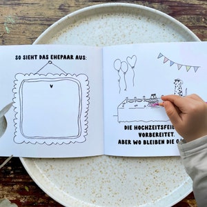May include: A black and white illustrated book with a blank frame on the left page and a scene of a wedding celebration with two dogs on the right page. The text on the right page reads "SO SIEHT DAS EHEPAAR AUS:" and "DIE HOCHZEITSFEIL VORBEREITET. ABER WO BLEIBEN DIE G".