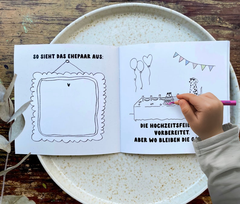 May include: A black and white illustrated book with a blank frame on the left page and a scene of a wedding celebration with two dogs on the right page. The text on the right page reads "SO SIEHT DAS EHEPAAR AUS:" and "DIE HOCHZEITSFEIL VORBEREITET. ABER WO BLEIBEN DIE G".