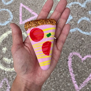 Op de afbeelding: Een roze en gele pizzapunt met rode en groene toppings, versierd met gouden glitter.
