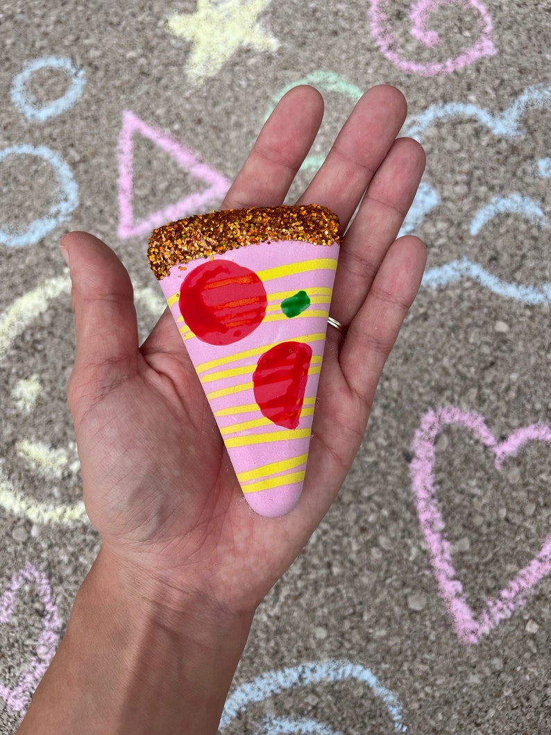 Op de afbeelding: Een roze en gele pizzapunt met rode en groene toppings, versierd met gouden glitter.