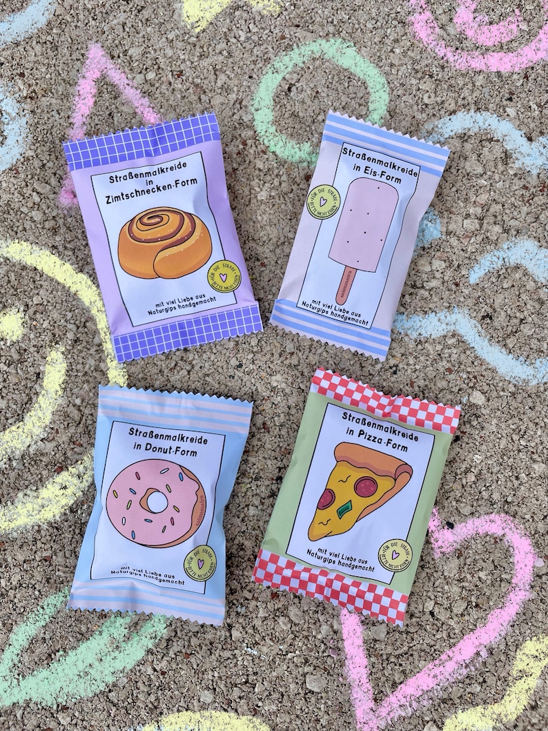 Op de afbeelding: Vier kleurrijke pakjes met stoepkrijt in de vorm van verschillende voedingsmiddelen. De pakjes zijn gelabeld met "Stra&szlig;enmalkreide", wat Duits is voor stoepkrijt. De voedselvormen zijn een kaneelrol, een ijslolly, een donut en een pizza.
