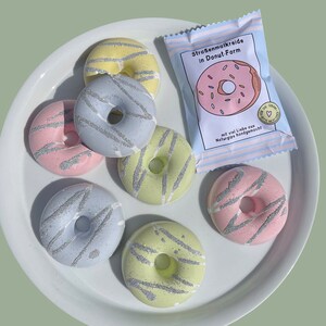 May include: Six pastel-colored donut-shaped sidewalk chalk pieces on a white plate. The chalk is decorated with silver glitter. A package of sidewalk chalk in the shape of a donut is in the background. The package says "Stra&szlig;enmalkreide in Donut-Form" and "mit viel Liebe aus Naturgips handgemacht!"