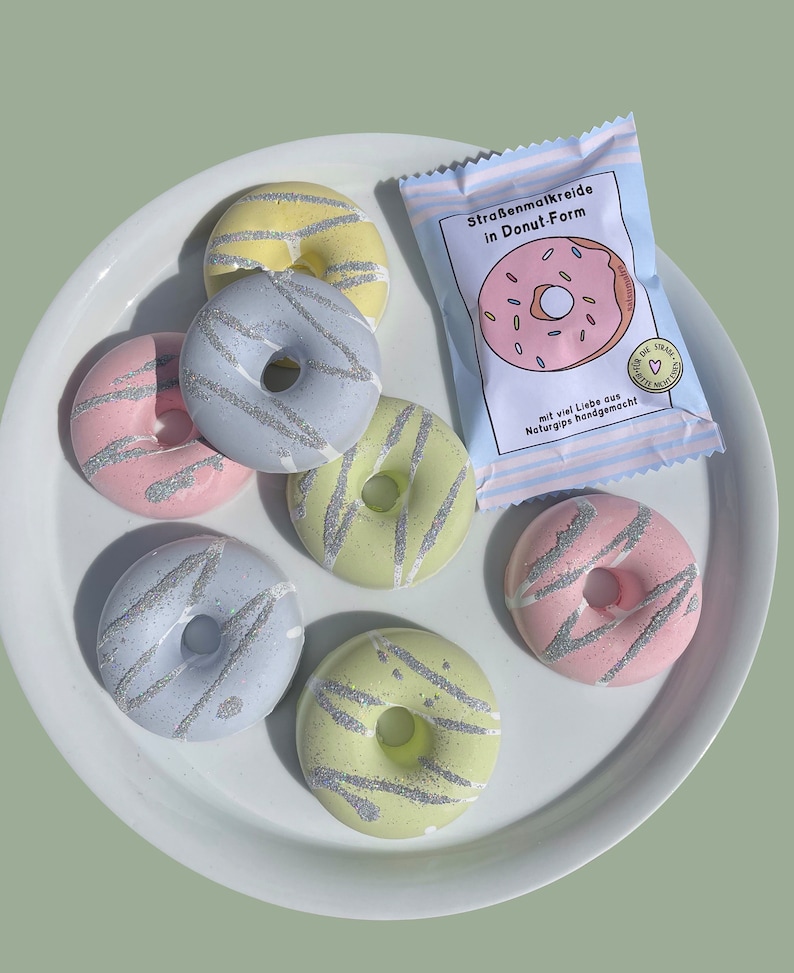 May include: Six pastel-colored donut-shaped sidewalk chalk pieces on a white plate. The chalk is decorated with silver glitter. A package of sidewalk chalk in the shape of a donut is in the background. The package says "Stra&szlig;enmalkreide in Donut-Form" and "mit viel Liebe aus Naturgips handgemacht!"