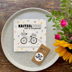 May include: A small brown box with a white sticker that says "Mal die Welt Bunt an!"  The box contains colored pencils.  A white card with colorful dots and a black and white illustration of a bicycle with flowers and a banner that says "Kritzelbuch Hurra, es wird geheiratet!"  The card is on a white plate.