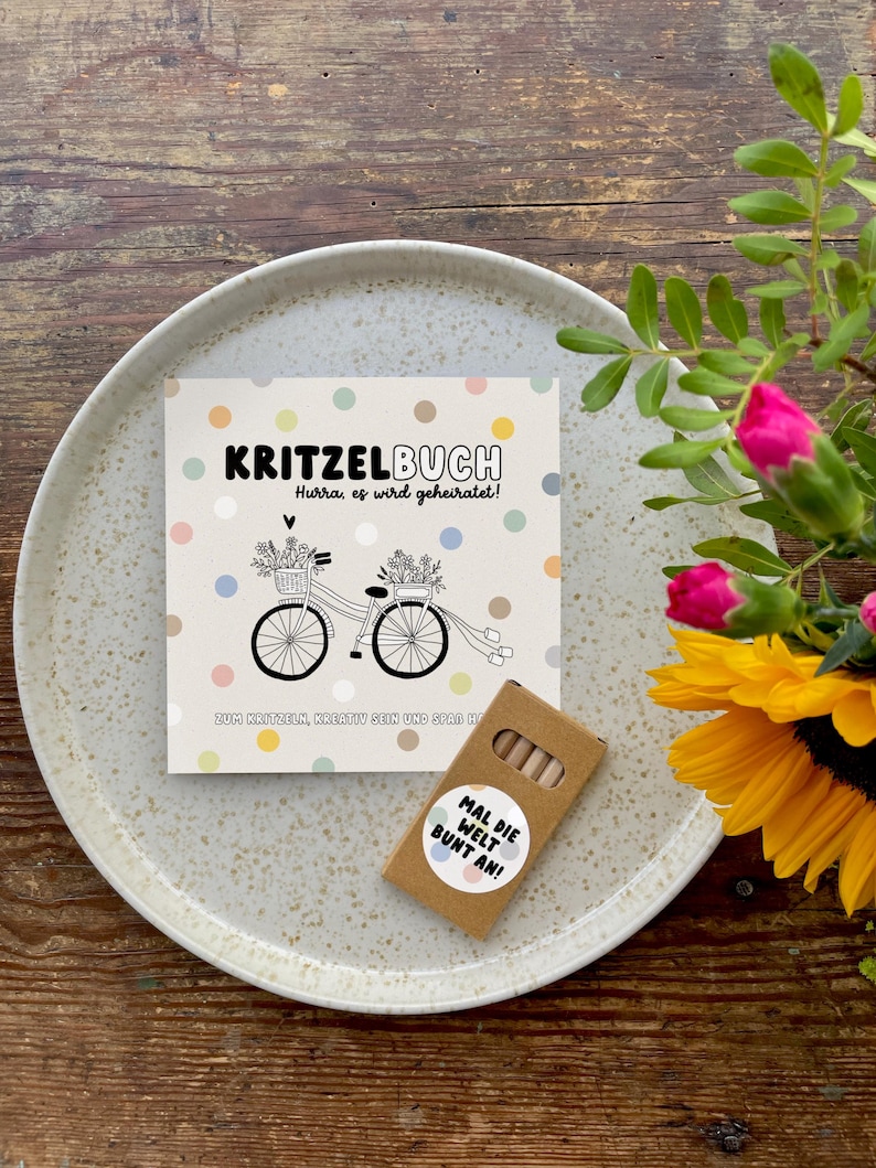 May include: A small brown box with a white sticker that says "Mal die Welt Bunt an!"  The box contains colored pencils.  A white card with colorful dots and a black and white illustration of a bicycle with flowers and a banner that says "Kritzelbuch Hurra, es wird geheiratet!"  The card is on a white plate.