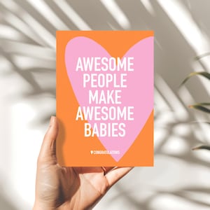May include: A greeting card with an orange background and a pink heart shape. The card reads "AWESOME PEOPLE MAKE AWESOME BABIES" in white text. The word "CONGRATULATIONS" is at the bottom. The card is held by a hand.