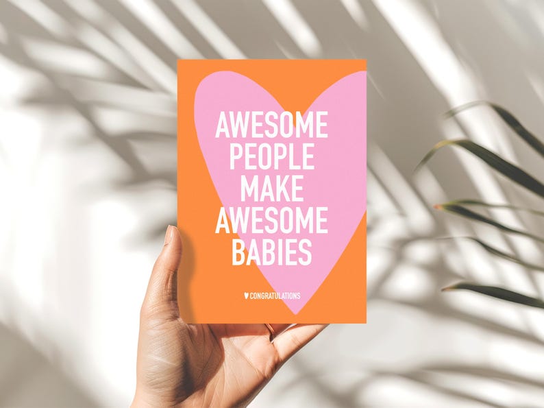 May include: A greeting card with an orange background and a pink heart shape. The card reads "AWESOME PEOPLE MAKE AWESOME BABIES" in white text. The word "CONGRATULATIONS" is at the bottom. The card is held by a hand.