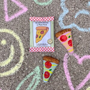 Op de afbeelding: Een pakje stoepkrijt in de vorm van pizzapunten. Het pakje is rood-wit geruit met de tekst "Stra&szlig;enmalKreide in Pizza-Form" en "mit viel Liebe aus Naturgips handgemacht". Er liggen twee glitter-bedekte pizzapunten op een grijs oppervlak met krijttekeningen van een driehoek, cirkel, hart en een halve maan.