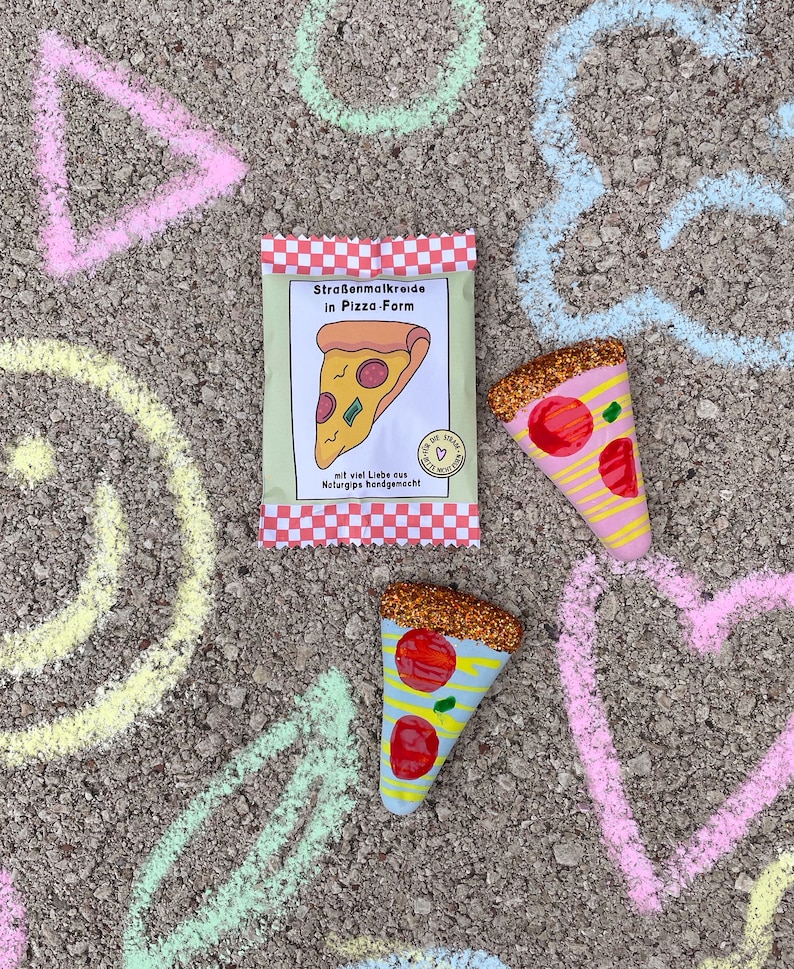 Op de afbeelding: Een pakje stoepkrijt in de vorm van pizzapunten. Het pakje is rood-wit geruit met de tekst "Stra&szlig;enmalKreide in Pizza-Form" en "mit viel Liebe aus Naturgips handgemacht". Er liggen twee glitter-bedekte pizzapunten op een grijs oppervlak met krijttekeningen van een driehoek, cirkel, hart en een halve maan.