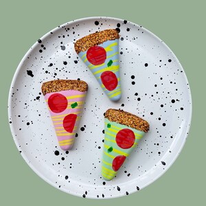 Op de afbeelding: Drie keramische pizza-punt ornamenten op een wit bord met zwarte stippen. De pizzapunten zijn versierd met glitter en hebben verschillende gekleurde korsten en toppings.
