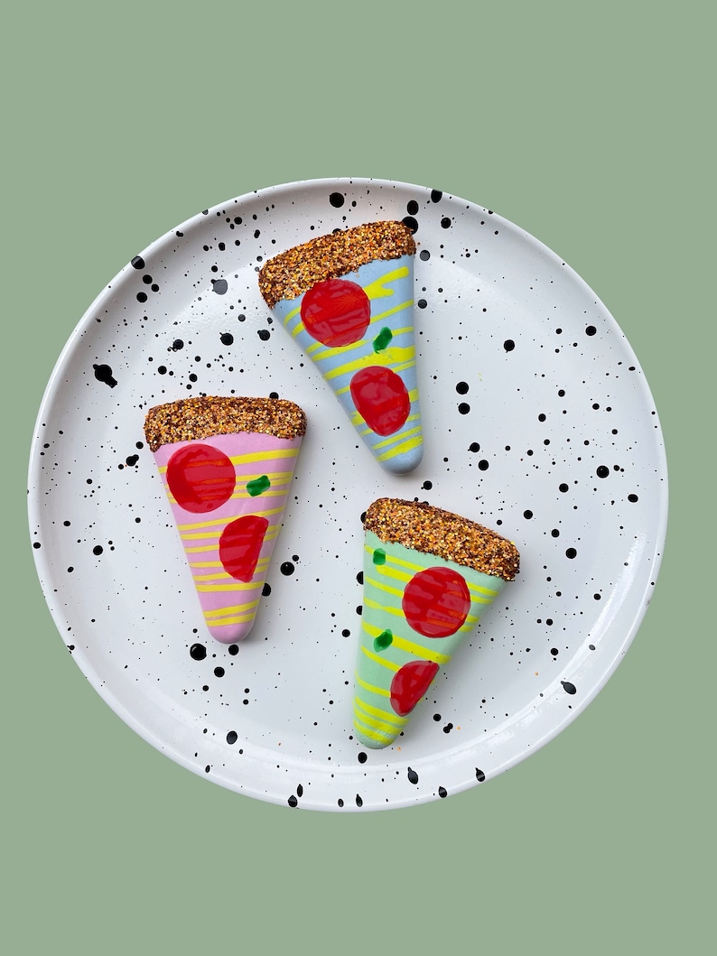 Op de afbeelding: Drie keramische pizza-punt ornamenten op een wit bord met zwarte stippen. De pizzapunten zijn versierd met glitter en hebben verschillende gekleurde korsten en toppings.