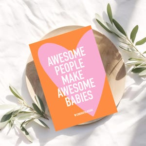 May include: A greeting card with the text "AWESOME PEOPLE MAKE AWESOME BABIES" in white on a pink heart shape, set against an orange background. The card is on a wooden plate, with green leaves around it. The word "CONGRATULATIONS" is at the bottom.