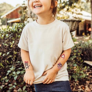 May include: A young person wearing a white t-shirt with three temporary tattoos on their arms. The tattoos are a heart, a cupcake, and a cat wearing a bow.
