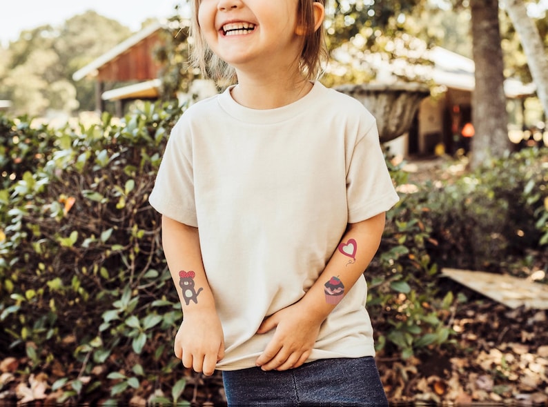 May include: A young person wearing a white t-shirt with three temporary tattoos on their arms. The tattoos are a heart, a cupcake, and a cat wearing a bow.