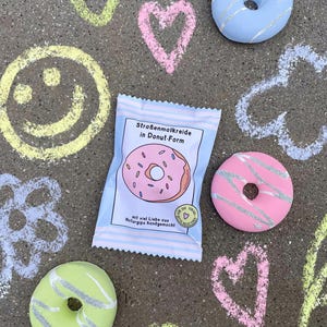 May include: A package of donut-shaped sidewalk chalk in blue packaging with a pink and white donut graphic. The text on the package reads "Stra&szlig;enmalkreide in Donut-Form" and "mit viel Liebe aus Naturgips handgemacht".
