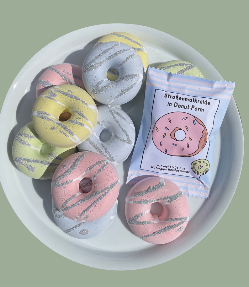 May include: A set of colorful donut-shaped sidewalk chalk in a variety of pastel colors with silver glitter. The chalk is packaged in a clear plastic bag with a label that reads "Stra&szlig;enmalkreide in Donut-Form" and "mit viel Liebe aus Naturgips handgemacht".