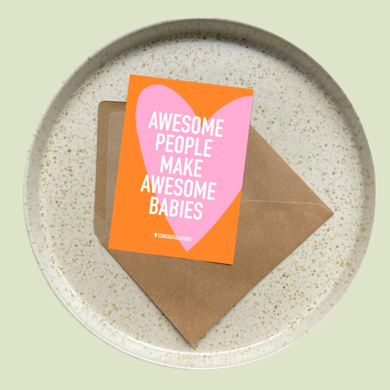 May include: A greeting card with the text "AWESOME PEOPLE MAKE AWESOME BABIES" in white on an orange background with a pink heart. The card is inside a brown envelope, resting on a speckled ceramic plate.