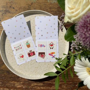 May include: A set of eight temporary tattoos with cute designs, including a panda, a cupcake, a cat, and a camera. The tattoos are on a white card with a pink and blue polka dot pattern. The card is folded in half and says "With Love" on the front.