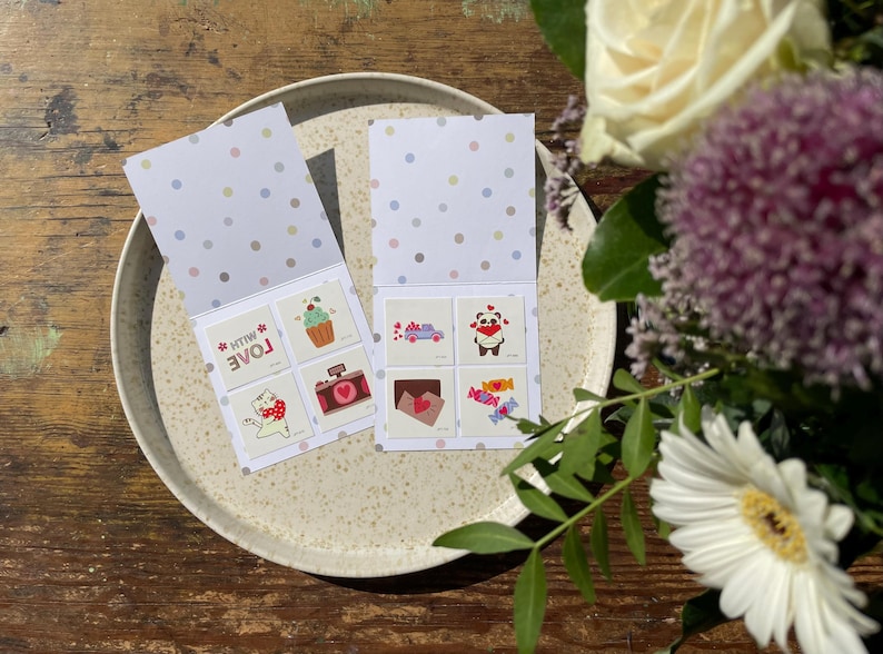 May include: A set of eight temporary tattoos with cute designs, including a panda, a cupcake, a cat, and a camera. The tattoos are on a white card with a pink and blue polka dot pattern. The card is folded in half and says "With Love" on the front.