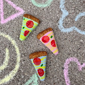 Op de afbeelding: Drie beschilderde pizzapunten op een grijs, getextureerd oppervlak. De pizzapunten zijn versierd met glitter, rode en gele strepen en rode en groene pepperoni. Het oppervlak is versierd met krijttekeningen van een cirkel, een driehoek en een wolk.