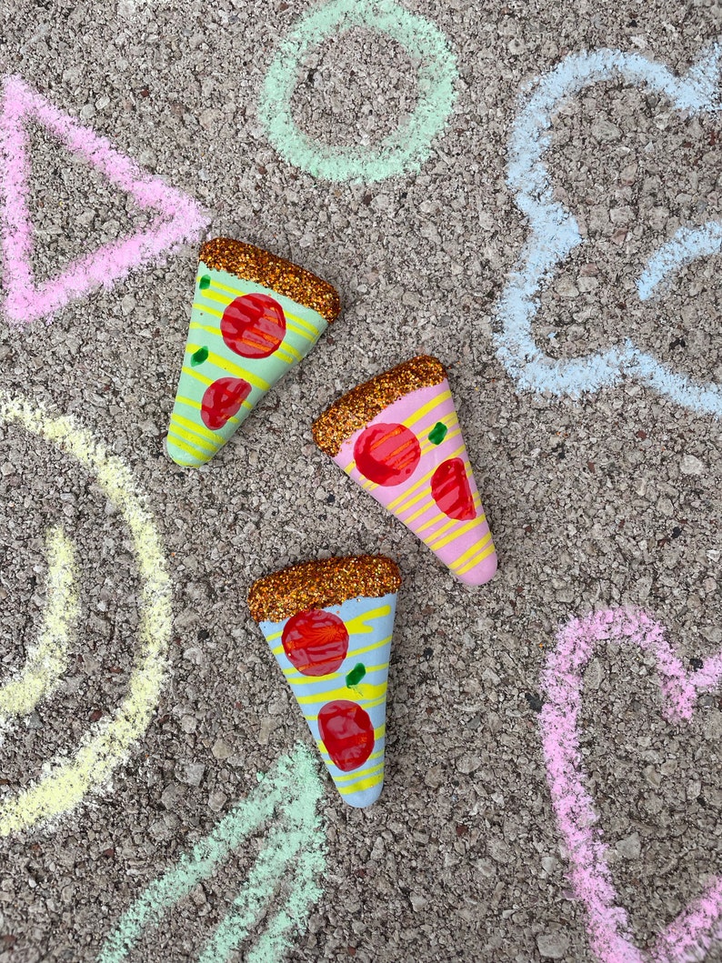 Op de afbeelding: Drie beschilderde pizzapunten op een grijs, getextureerd oppervlak. De pizzapunten zijn versierd met glitter, rode en gele strepen en rode en groene pepperoni. Het oppervlak is versierd met krijttekeningen van een cirkel, een driehoek en een wolk.