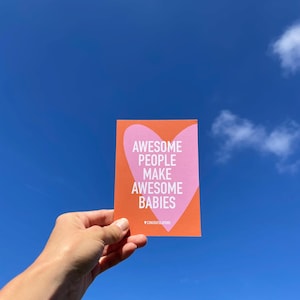May include: A greeting card with the text "AWESOME PEOPLE MAKE AWESOME BABIES" inside a pink heart on an orange background. The card also has the word "CONGRATULATIONS" at the bottom. The card is held against a bright blue sky.