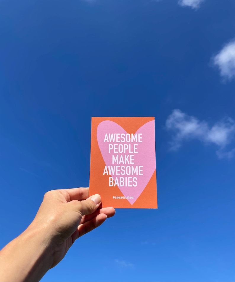 May include: A greeting card with the text "AWESOME PEOPLE MAKE AWESOME BABIES" inside a pink heart on an orange background. The card also has the word "CONGRATULATIONS" at the bottom. The card is held against a bright blue sky.