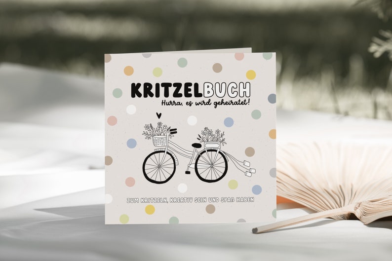 May include: A white card with a light gray background and colorful polka dots. The card says "KRITZELBUCH" in black bold letters. Below the title, it says "Hurra, es wird geheiratet!" in black letters. A black and white illustration of a bicycle with flowers in baskets on the handlebars and a white ribbon tied to the back is in the center of the card. Below the bicycle, it says "ZUM KRITZELN, KREATIV SEIN UND SPASS HABEN" in black letters.