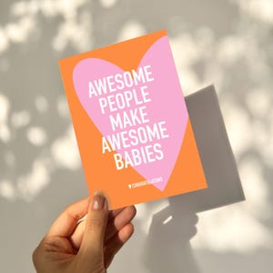 May include: A greeting card with an orange background and a pink heart shape. The card reads "AWESOME PEOPLE MAKE AWESOME BABIES" in white text. The word "CONGRATULATIONS" is at the bottom. The card is held in a hand.