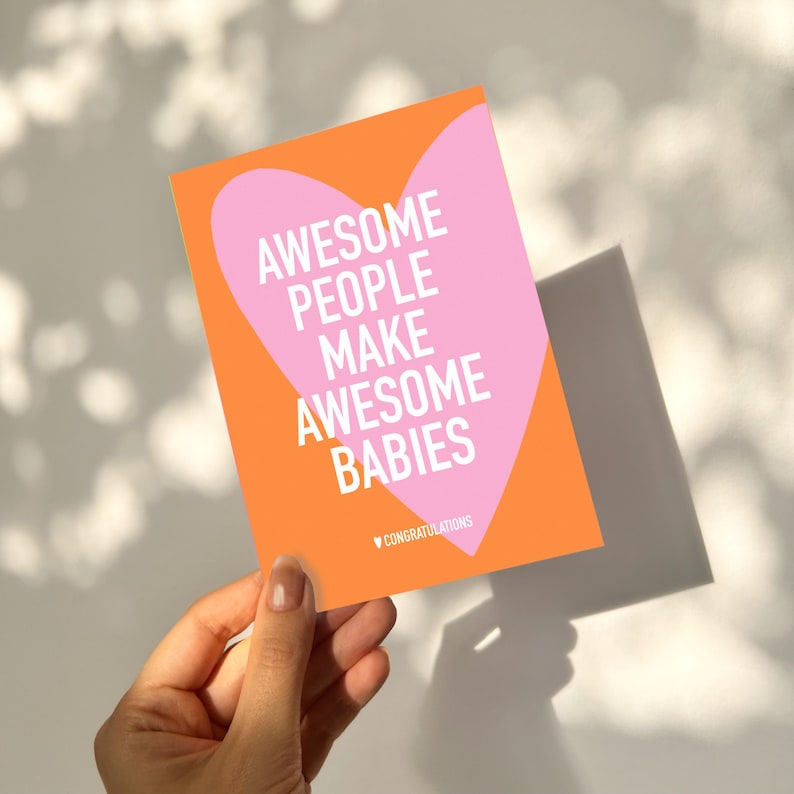 May include: A greeting card with an orange background and a pink heart shape. The card reads "AWESOME PEOPLE MAKE AWESOME BABIES" in white text. The word "CONGRATULATIONS" is at the bottom. The card is held in a hand.