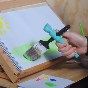 Può includere: Una persona sta dipingendo un campo verde su una tela bianca con un pennello blu e nero. La tela è su un cavalletto. C'è un sole giallo nell'angolo in alto a sinistra della tela.