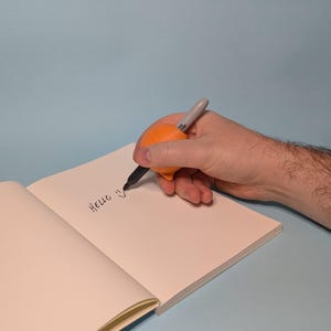 May include: An orange pen grip is used to write the word "HELLO" on a blank, open notebook. The pen is a Sharpie brand. The background is a solid light blue.